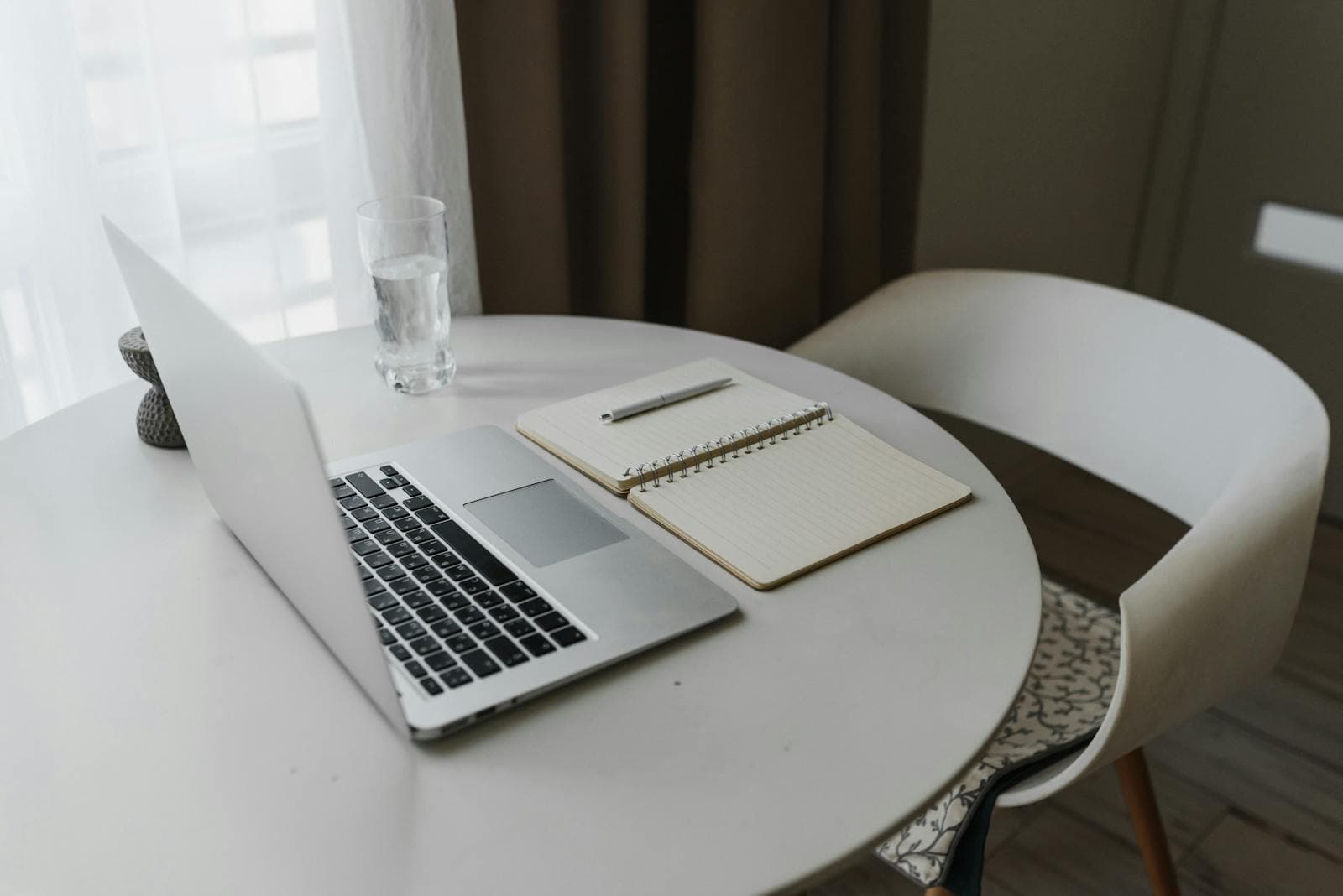 An open laptop and a notebook with a pen on a sunlit kitchen table, suggesting a person quietly working through household finances